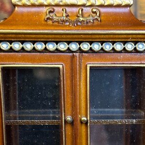 May include: A wooden cabinet with glass doors and shelves. The cabinet has a decorative top with gold accents and a row of white beads around the top edge. The cabinet is painted with floral designs on the sides.