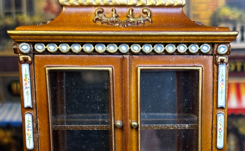 May include: A wooden cabinet with glass doors and shelves. The cabinet has a decorative top with gold accents and a row of white beads around the top edge. The cabinet is painted with floral designs on the sides.