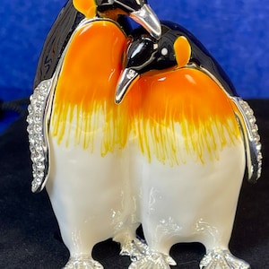 Puede incluir: Dos pingüinos hechos de esmalte blanco, naranja y negro con pies plateados y un detalle de pedrería en la parte posterior de un pingüino. Los pingüinos se miran y parecen estar abrazándose.