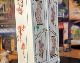 May include: White painted wooden armoire with ornate gold trim and floral decals. The armoire has two doors and sits on four legs.