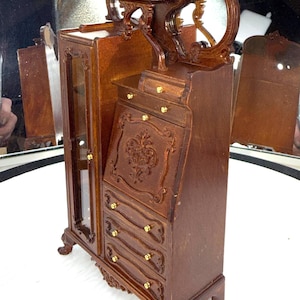 May include: A miniature wooden cabinet with a glass-door display case, drawers, and a decorative top. The cabinet is brown with gold-colored hardware and ornate carvings. The piece is likely a dollhouse or miniature furniture item.