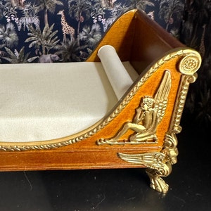 May include: A wooden chaise lounge with gold accents and a white fabric cushion. The chaise has a carved design with a winged figure on the side.