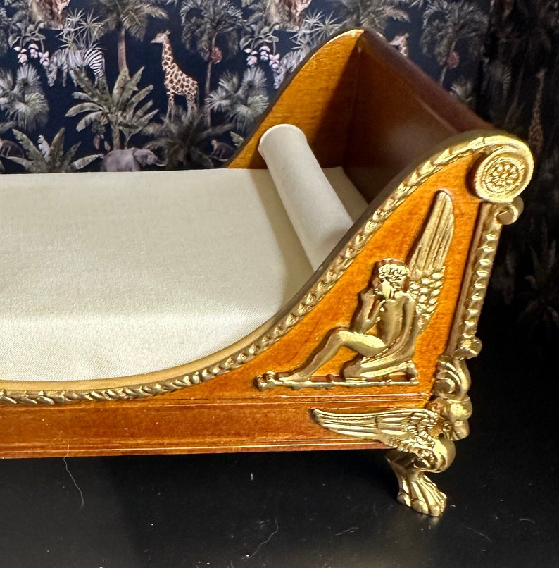 May include: A wooden chaise lounge with gold accents and a white fabric cushion. The chaise has a carved design with a winged figure on the side.