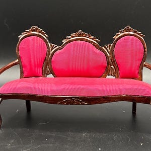 May include: Miniature antique-style sofa with a dark wood frame and vibrant pink upholstery. The sofa features ornate detailing on the back and arms, with a classic, elegant design. A decorative piece for dollhouses or miniature displays.