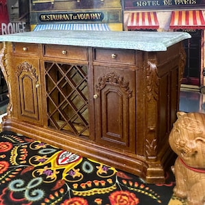 May include: A miniature wooden kitchen island with a gray countertop. The island has drawers, cabinet doors, and a wine rack. A carved wooden tiger figurine with a red collar sits nearby. The island is on a colorful patterned rug.