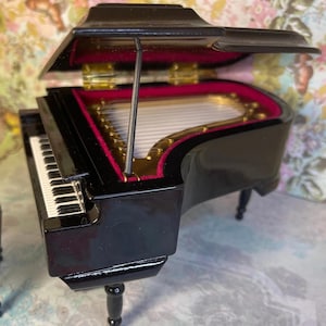 May include: A miniature black grand piano with a glossy finish. The piano's interior is lined with red velvet and gold accents. The keys are white and black. The piano has three black legs and a partially open lid.