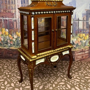 May include: A wooden display cabinet with glass doors and ornate gold trim. The cabinet has a decorative floral design and sits on four carved legs.