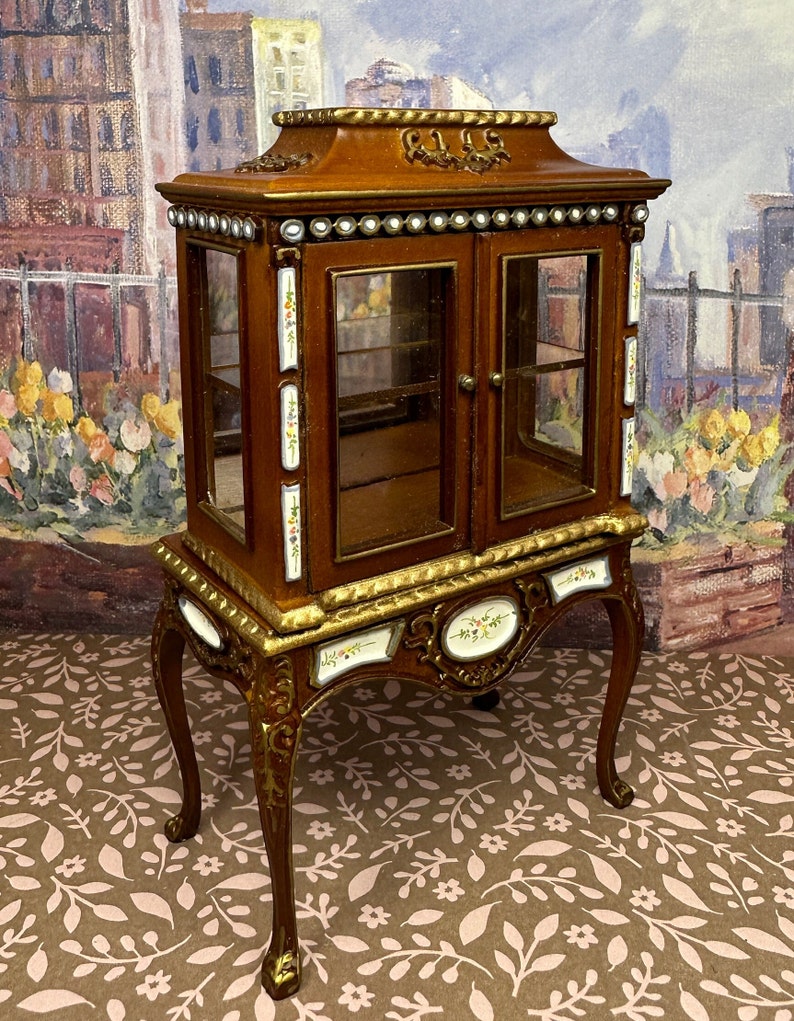 May include: A wooden display cabinet with glass doors and ornate gold trim. The cabinet has a decorative floral design and sits on four carved legs.