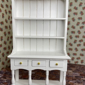 May include: White wooden hutch with three drawers and a lower shelf. The hutch has a scalloped top and two shelves. The drawers have gold knobs.