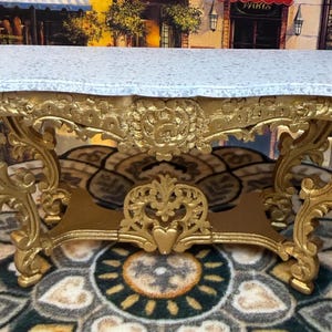 May include: A small, ornate gold-colored console table with a white and gray speckled top. The table features intricate carvings and a decorative base, suitable for display or decor.