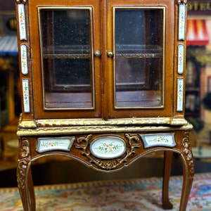 May include: A miniature wooden cabinet with glass doors and ornate details. The cabinet has a brown finish with gold accents and floral designs. It sits on four legs and has a shelf inside.