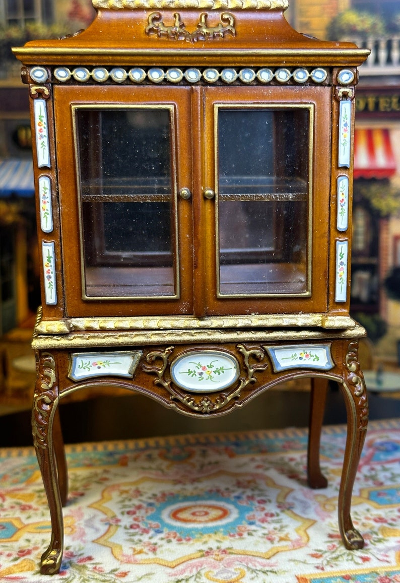 May include: A miniature wooden cabinet with glass doors and ornate details. The cabinet has a brown finish with gold accents and floral designs. It sits on four legs and has a shelf inside.