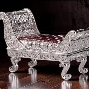 May include: A silver-colored miniature chaise lounge with ornate detailing. The seat cushion is a deep red with a button-tufted design. The legs are curved, and the back is slightly reclined, creating a regal appearance.
