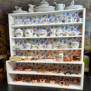 Puede incluir: Una estantería de madera blanca con cinco estantes, cada uno forrado con papel tapiz floral. Los estantes están llenos de tazas de té, platillos, platos y platos para servir de porcelana en miniatura. Los dos estantes inferiores están llenos de ollas y sartenes de cobre en miniatura.