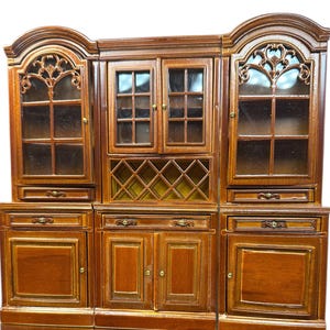 Könnte beinhalten: Ein detaillierter Holzschrank mit einer satten, braunen Oberfläche. Der Schrank verfügt über Türen mit Glasfront, dekorative Schnitzereien und ein Weinregal. Das Design umfasst mehrere Staufächer und Schubladen und zeigt einen klassischen, eleganten Stil.