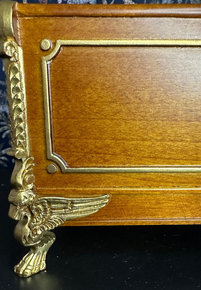 May include: A wooden box with gold accents and decorative feet. The box has a gold trim around the top and bottom edges. The feet are shaped like wings and have a gold finish.