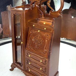 May include: A miniature wooden cabinet with a mirrored top and a writing desk. The cabinet has a glass-fronted door, drawers, and ornate carvings. The wood is a rich brown color with gold-colored hardware.
