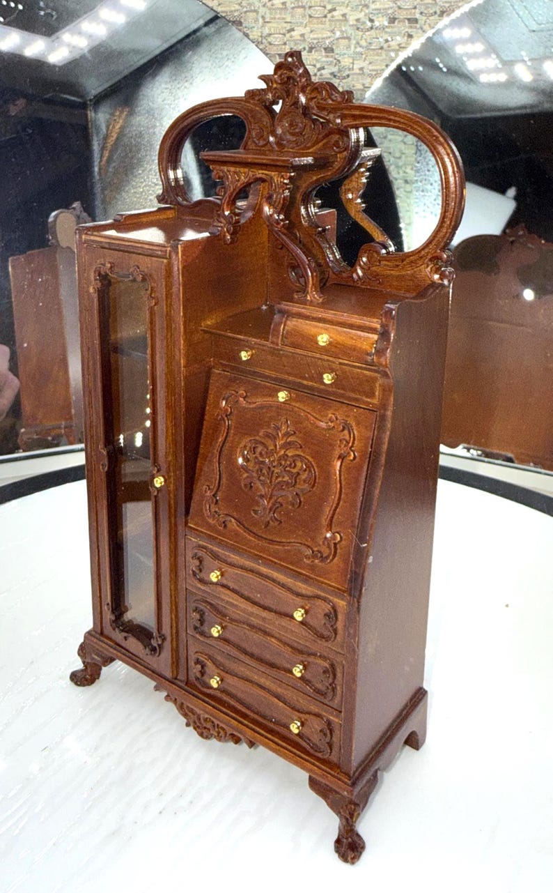 May include: A miniature wooden cabinet with a mirrored top and a writing desk. The cabinet has a glass-fronted door, drawers, and ornate carvings. The wood is a rich brown color with gold-colored hardware.