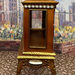 May include: A wooden display cabinet with a glass front and gold accents. The cabinet is on a stand with ornate legs and a floral design on the front.