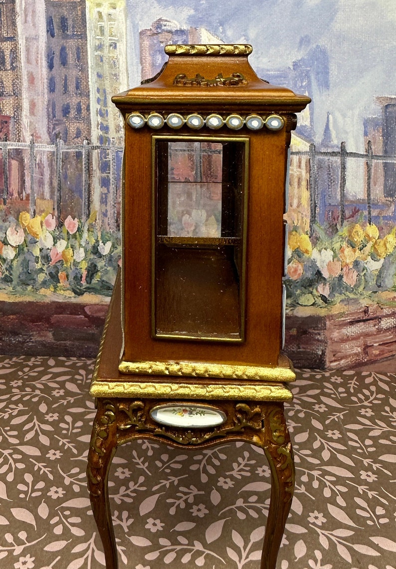 May include: A wooden display cabinet with a glass front and gold accents. The cabinet is on a stand with ornate legs and a floral design on the front.
