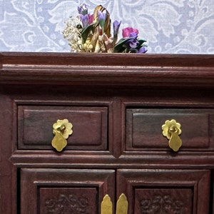 May include: A dark red wooden cabinet with two drawers and two doors. The cabinet has gold-colored handles and is decorated with intricate carvings. The cabinet is sitting on a white patterned surface.