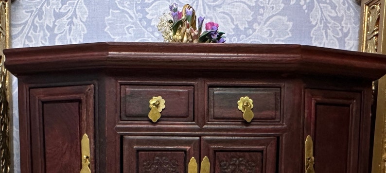 May include: A dark red wooden cabinet with two drawers and two doors. The cabinet has gold-colored handles and is decorated with intricate carvings. The cabinet is sitting on a white patterned surface.