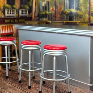 May include: A silver bar counter with a silver trim and four red bar stools. The bar counter is in a miniature setting with a backdrop of a Parisian street scene.