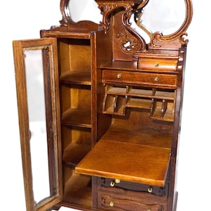 May include: A miniature wooden cabinet with a mirrored top and a drop-down desk. The cabinet has a glass-paned door, shelves, and drawers. The wood is a warm brown color, and the cabinet has ornate carvings.