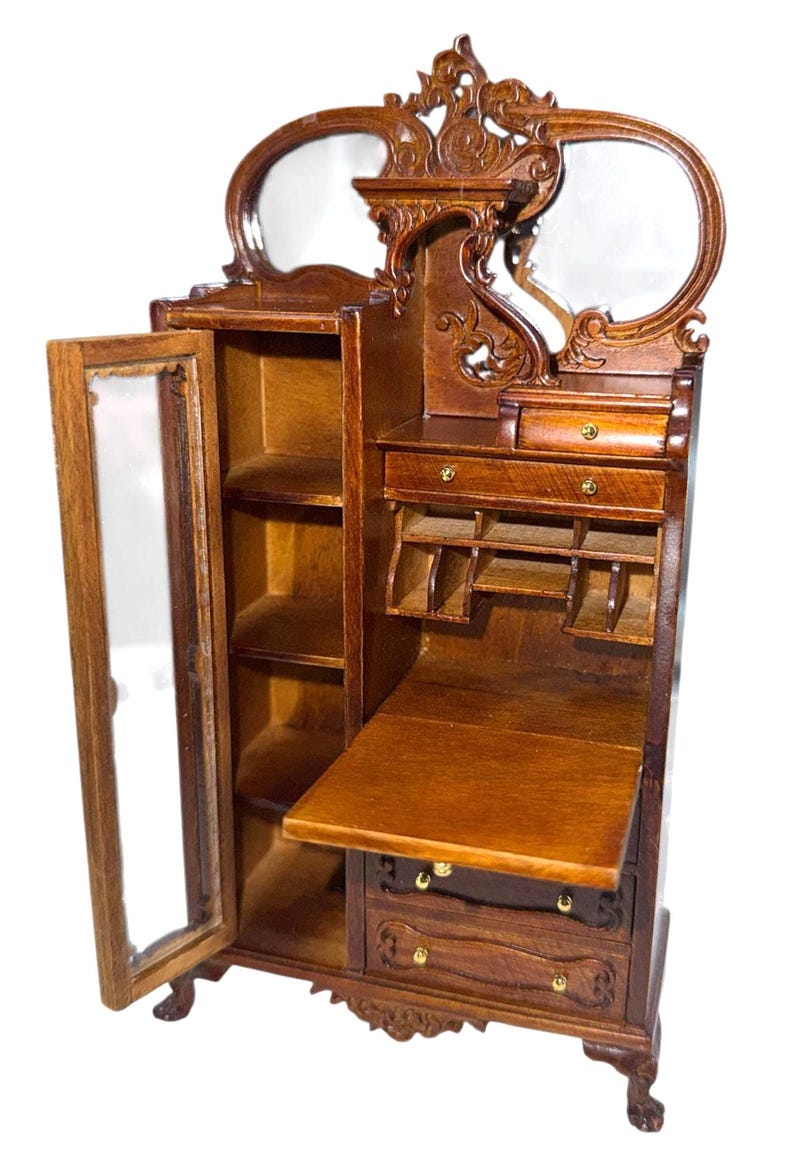 May include: A miniature wooden cabinet with a mirrored top and a drop-down desk. The cabinet has a glass-paned door, shelves, and drawers. The wood is a warm brown color, and the cabinet has ornate carvings.