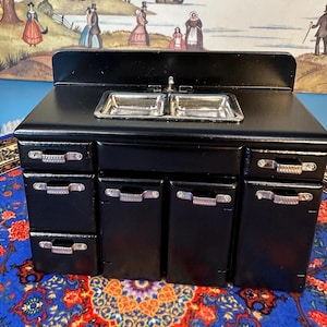 May include: Black dollhouse kitchen sink and cabinet set with silver handles. The sink has two basins and a chrome faucet. The cabinets have three drawers and two doors.