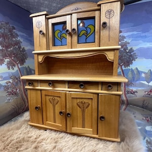 Könnte beinhalten: Ein Holzschrank mit zwei Abschnitten. Der obere Abschnitt hat zwei Türen mit Buntglasscheiben mit gelben und blauen Blumenmustern. Der untere Abschnitt hat zwei Türen und drei Schubladen. Der Schrank ist aus hellem Holz gefertigt.