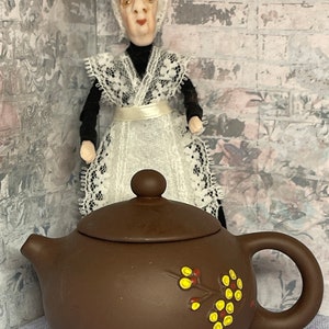 Puede incluir: Una tetera marrón con tapa y un diseño floral. La tetera tiene un diseño floral amarillo y rojo en el lateral. La tetera está sobre una superficie con un patrón azul y blanco.