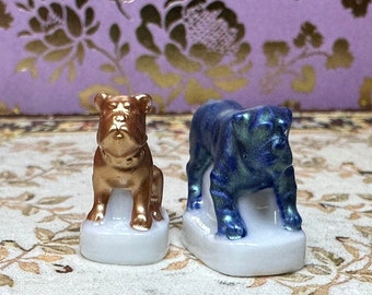 May include: Two ceramic bulldog figurines. One is gold-coloured and sitting, the other is blue and standing. Both are on white bases. The background is a floral pattern in gold and purple.