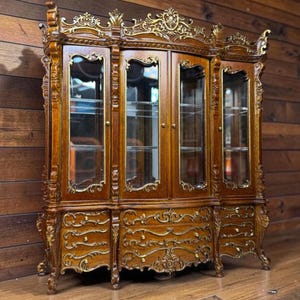 May include: A detailed, wooden display cabinet with glass doors and ornate gold accents. The cabinet features multiple shelves and drawers, showcasing a classic, antique design. The wood has a rich, warm tone, and the gold detailing adds a touch of elegance.