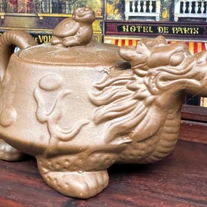 May include: A brown ceramic teapot shaped like a dragon with a lid featuring a small turtle. The teapot has a spout shaped like the dragon's head and a handle on the side.