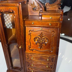 May include: A miniature wooden vanity with a mirror, drawers, and a cabinet. The vanity is made of dark brown wood with ornate carvings and gold-colored hardware. The cabinet door has a glass panel.