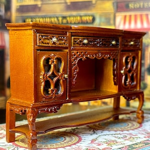 May include: A miniature wooden sideboard with intricate carvings and glass doors. The sideboard has a lower shelf and sits on four carved legs.