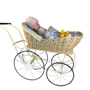 Op de afbeelding: Een vintage rieten kinderwagen met een pop erin. De wagen heeft een crèmekleurig frame met zwart omrande wielen. De pop draagt een roze en witte gebreide muts en is bedekt met een blauw-witte deken. Gele badeendjes liggen in de wagen.