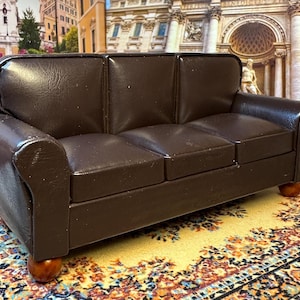 Puede incluir: Un sofá de cuero marrón en miniatura con tres cojines de asiento y reposabrazos redondeados. El sofá se apoya sobre cuatro pequeñas patas redondas de madera. El sofá está colocado sobre una alfombra estampada con diseños amarillos y florales.