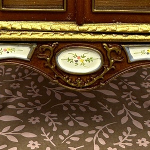 May include: A wooden cabinet with gold trim and painted floral details. The cabinet has a glass front and sits on four ornate legs.
