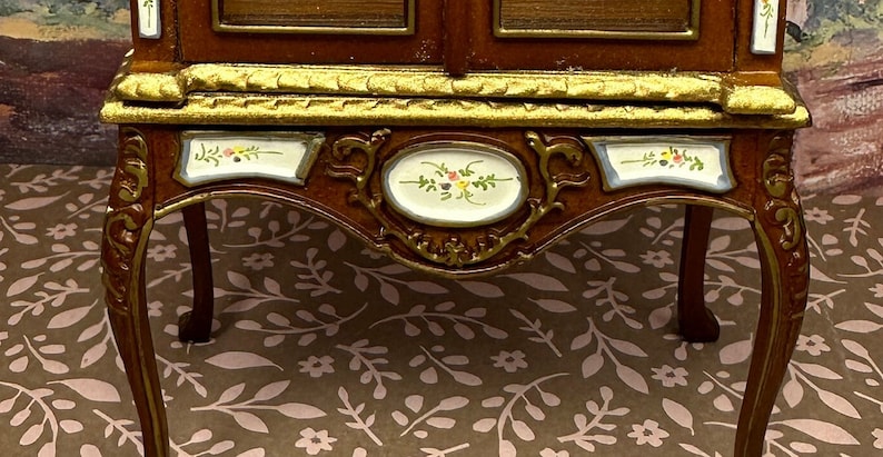 May include: A wooden cabinet with gold trim and painted floral details. The cabinet has a glass front and sits on four ornate legs.