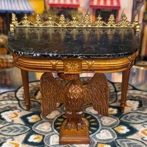 May include: A miniature, ornate console table with a black marble top and a carved wooden eagle base. The table features gold accents along the top edge and a detailed floral carving. The table is approximately 76 cm long.