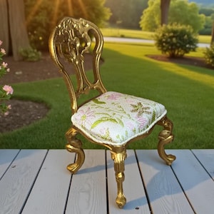 May include: A miniature gold-colored chair with ornate detailing. The chair has a floral patterned seat cushion in white, pink, and green. The chair's legs and back are intricately designed, showcasing a vintage aesthetic. The chair is placed on a wooden surface.