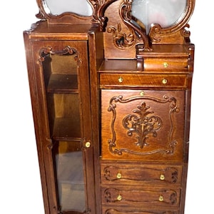 May include: A miniature wooden dresser with a mirror, drawers, and a glass-door cabinet. The dresser is brown with ornate carvings and gold-colored hardware. The mirror is oval-shaped and the cabinet has shelves. A decorative piece for a dollhouse or miniature collection.