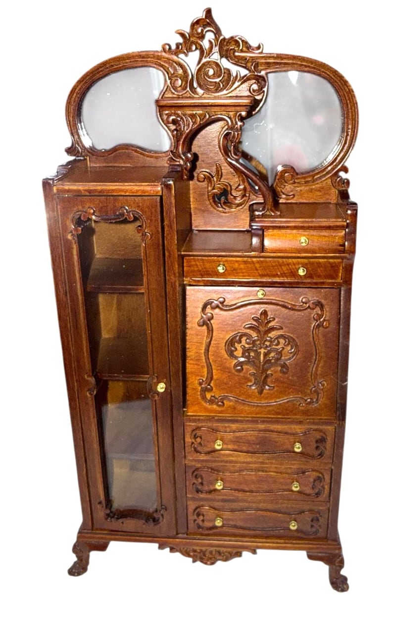May include: A miniature wooden dresser with a mirror, drawers, and a glass-door cabinet. The dresser is brown with ornate carvings and gold-colored hardware. The mirror is oval-shaped and the cabinet has shelves. A decorative piece for a dollhouse or miniature collection.