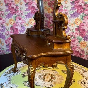 May include: A miniature wooden vanity set with a gold-painted finish. The vanity has a mirror with an ornate frame, two drawers, and a top surface. The vanity is sitting on a round, floral-patterned rug.