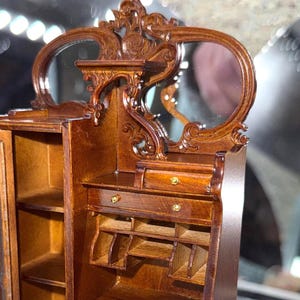May include: A miniature wooden cabinet with a mirror, featuring ornate carvings and a warm brown finish. The cabinet has a small drawer and open shelves, with gold-colored hardware. The mirror is oval-shaped and adds to the antique aesthetic.