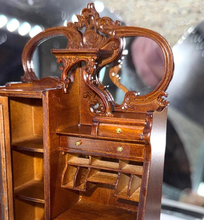 May include: A miniature wooden cabinet with a mirror, featuring ornate carvings and a warm brown finish. The cabinet has a small drawer and open shelves, with gold-colored hardware. The mirror is oval-shaped and adds to the antique aesthetic.