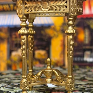 May include: A miniature gold-colored table with ornate detailing. The table has a round top, decorative legs, and an intricate base. The table is approximately 3 inches tall and is designed with a vintage aesthetic.