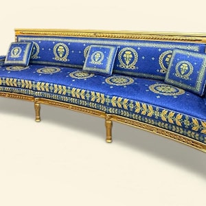 May include: A curved sofa with a gold-colored frame and legs. The blue upholstery is decorated with gold-colored floral and medallion patterns. Four matching square pillows are arranged on the sofa.